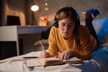 Serious student girl in glasses reading book on bed in dormitory, studying paper textbook, focused on text, preparing for exam, lesson, learning presentation
