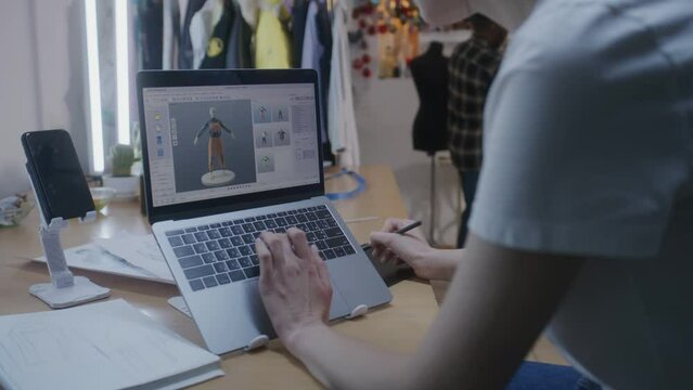 Female fashion designer draws future clothes in 3D modeling software on laptop using digital tablet computer and stylus. She works in atelier workshop. Concept of fashion and technologies in business.