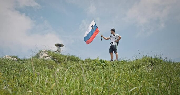 Camera tracking from low perspective up a hill a hiker at the top with a hiking pole in his hand and a slovenian flag attached on it and letting it flutter in the wind.