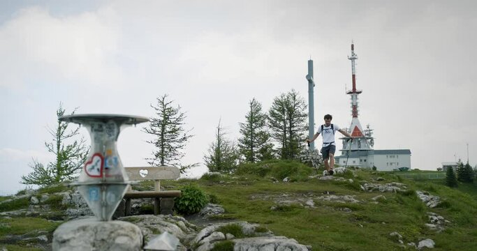 Hiker reaches top of the mountain and stops at the monument  and looks at it where the directions are written. Cloudy summer weather.