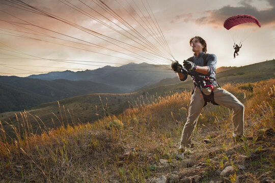 A speed flyer pulls his parasail in for a landing on a grassy hill at sunset.