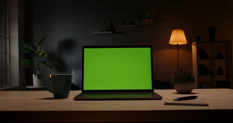 Chroma key green screen template - a laptop computer on a desk in cozy living room at night. Computer set for distance learning and remote work, technology concept 