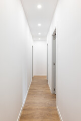 Long corridor with white smooth walls, bright light pours from spotlights from the ceiling, wooden laminate flooring, open doors on both sides to separate living rooms.