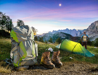 Hiker man standing next to Hiking Equipment, Tent, Backpack and Boots under a moon night sky at...