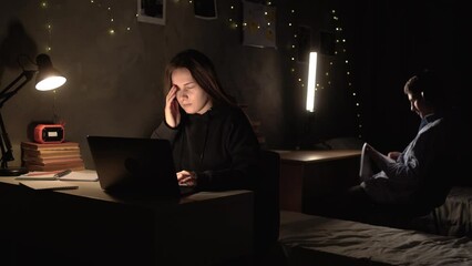 a young student girl sits in a college dorm room at night looking for information on the Internet using a laptop. Violent headache from overwork.