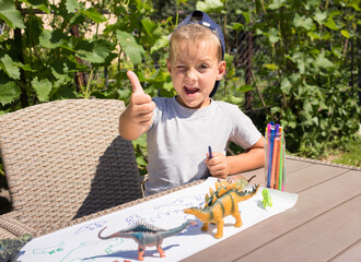 preschool boy draws toy dinosaurs with felt-tip pen while sitting at table in garden . Summer...