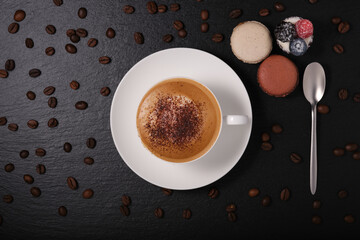Coffee with macaroon and a spoon on a black background. White cup with cappuccino, cheesecake with berry on a dark background flat lay. breakfast on a black background, coffee beans.