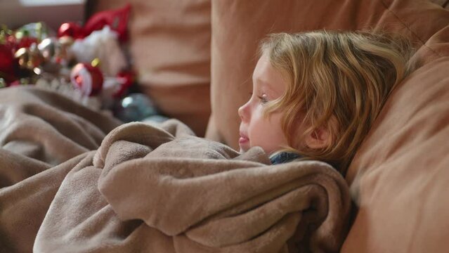 Close up of sick child resting on the sofa under blanket, struggling of fever, watching tv. Nice domestic interior. Corona symptoms, virus disease, concept of health, illness, common cold. HQ 4K