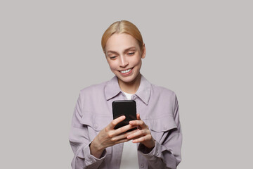 Fototapeta premium Cheerful young woman with blond hair holding smartphone on white studio wall background. People lifestyle concept. Model using mobile phone, typing sms message