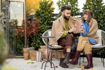 Young stylish couple warming up together, sitting covered with plaid by the fire and drink wine,...