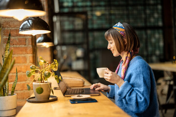 Young stylish woman works on laptop while sitting by the window with a coffee drink at modern cafe....