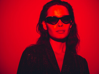 Young beautiful confident brunette female in trendy evening shiny green reflecting dress. Sexy carefree woman isolated on red neon light in studio. Fashionable model in sunglasses. Fantasy style