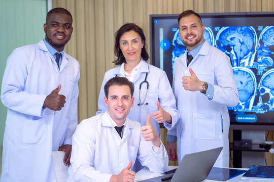 Group Of Happy Multicultural Doctors Showing Thumbs Up And Smiling On Camera While Posing At Conference Room. Portrait Of Multiracial Medical Specialists In White Lab Coats.