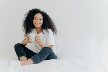 Young pretty African American woman rests in bed at home, drinks hot tea in morning, enjoys...