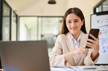 Charming millennial Asian businesswoman looking at laptop screen while using her phone