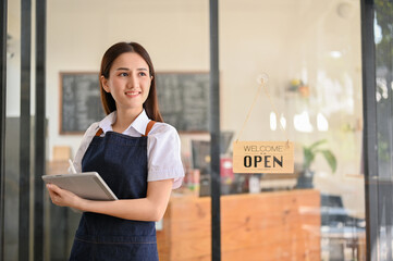 Charming young Asian female barista stands in front of the coffee shop's entrance door