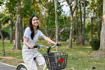 Joyful Asian woman riding a bicycle in the public park on the weekend, enjoys doing outdoor activity.