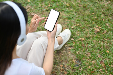 Young Asian woman sitting on grass, wearing headphones, relaxing in the public city park.
