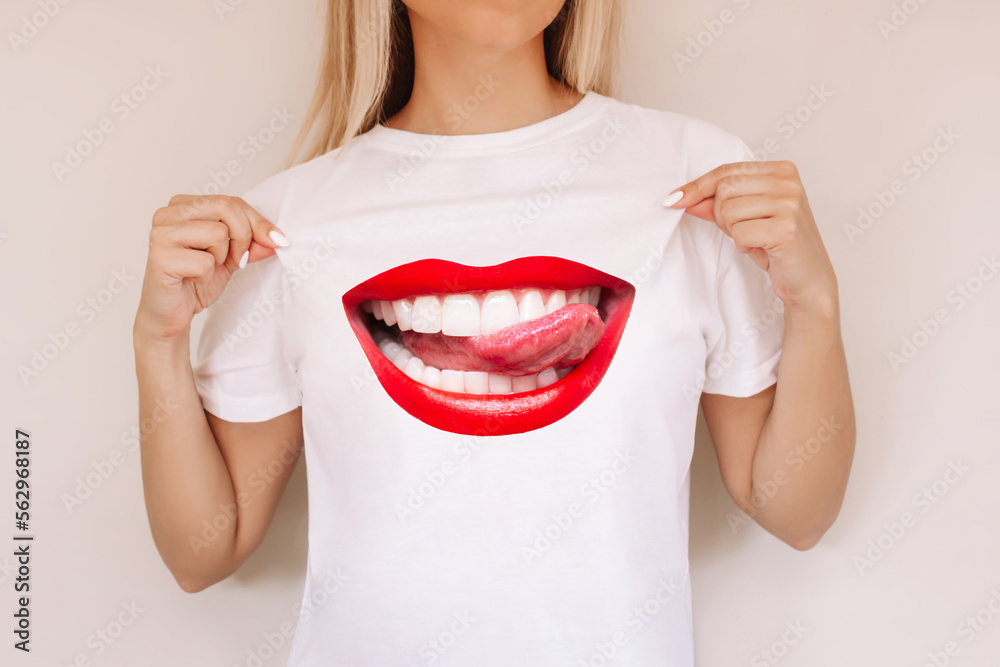 Wall mural Cropped shot of a young blonde woman demonstrating her white casual t-shirt with trendy creative design of big smiling female mouth with red lips showing tongue isolated on color beige background