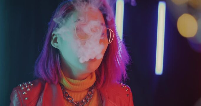 Portrait of pensive Asian lady slowly exhaling vaping smoke enjoying on futuristic neon background. People and relaxation habit concept.