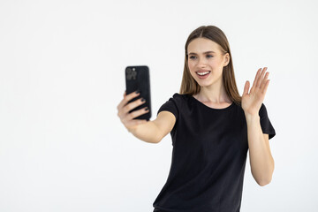 Happy young woman blogger influencer holding modern smart phone wave hand hello. Smiling vlogger girl looking at mobile make video call, shooting vlog taking selfie on grey background