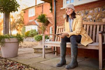 Woman Sitting On Bench Outside Home Wearing Boots Holding Mobile Phone Ready For Autumn Walk