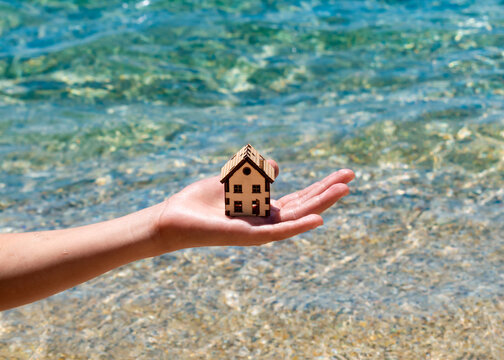 Boy carefull hold model of house above crystal clear water of sea. House in hand of child and summer sea beach. Family holiday property, resort, vacation real estate, save nature as your home