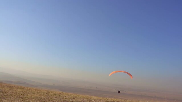 The pilot on a paraglider flew into the distance from a mountain peak. Above the paraglider is a blue cloudless sky and autumn fields.