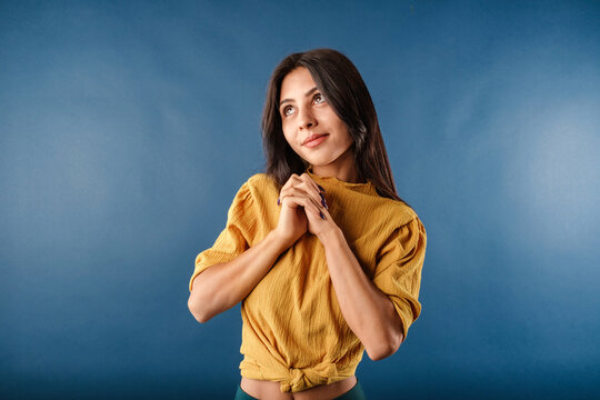 Brown-haired woman wearing mustard yellow t-shirt isolated over blue background imagining and thinking with her hands together. Looking to empty corner copy space.