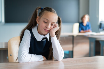 Little caucasian girl is bored at the lesson at school. The schoolgirl is sitting at her desk and the teacher is sitting in the background.