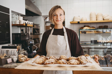 Welcoming female baker holding freshly baked almond croissants in  background of bakery shop. - 563002353
