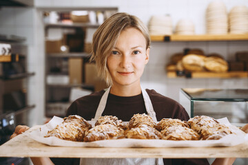 Welcoming female baker holding freshly baked almond croissants in  background of bakery shop.