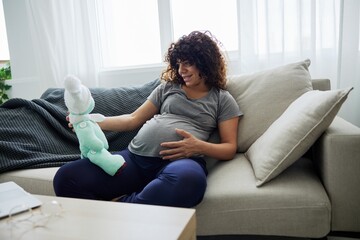 Obraz premium A pregnant woman sitting on the sofa with children's toy rabbit in her hands Easter, talking to the child, smiling. Lifestyle of a pregnant woman, preparation for childbirth, last month of pregnancy