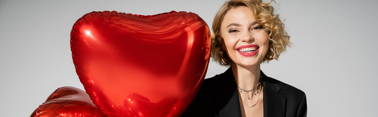 happy young woman in black blazer smiling near red heart-shaped balloons isolated on grey, banner.