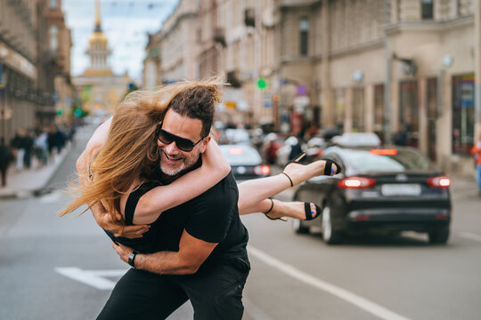 Excited Strong Tanned Spanish Man Lifts Up His Redhead Girlfriend Smiles Spinning Girl In Air. Young Caucasian Couple Embracing On Street. Romance, Dating. Valentines Day. Guy In Glasses Hugs Wife.