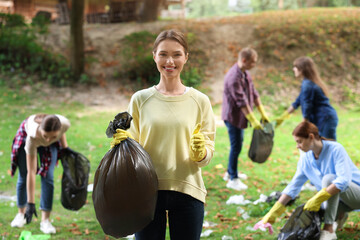 Young woman with plastic bag showing thumb up and group of people collecting garbage in park