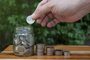 hand of a man saving coin in a jar, and coin on wooden table with green background, symbol of money saving for retirement