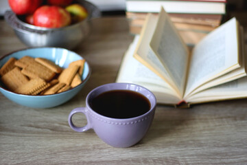 Stack of books, e-reader, reading glasses, bowl of biscuits, apples and cup of tea on the table. Bookshelf in the background. Selective focus.