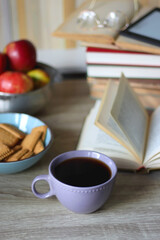 Stack of books, e-reader, reading glasses, bowl of biscuits, apples and cup of tea on the table. Bookshelf in the background. Selective focus.
