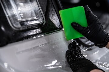 Employee of a car detailing studio applies a colorless protective film covering the paintwork of a white car. The film protects the car paint against UV rays and scratches