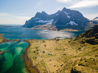 Aerial drone view of Vindstad village in Lofoten, Norway, small colored houses, big mountains and a bluish lake, during spring on a clear day with clouds