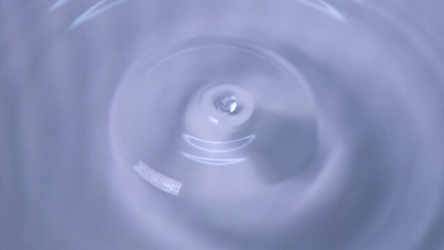 A drop of water falls forming circles on the surface of the water against a pale blue background