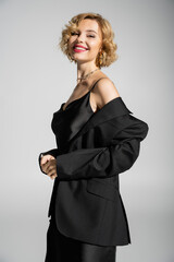 Fototapeta premium cheerful young woman in black blazer and slip dress looking at camera isolated on grey.