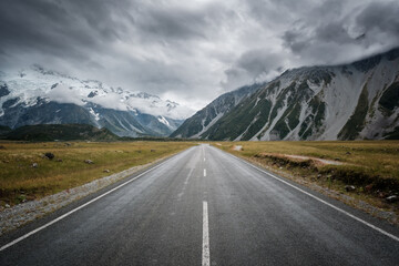 Hooker Track road near Mount Cook in the South Island of New Zealand leading towards snow capped mountains with dark clouds overhead