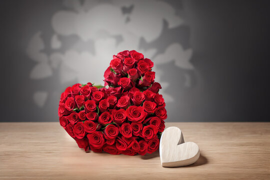Photo of a carved heart made of wood next to a bouquet of red roses on a light grey wooden table surface