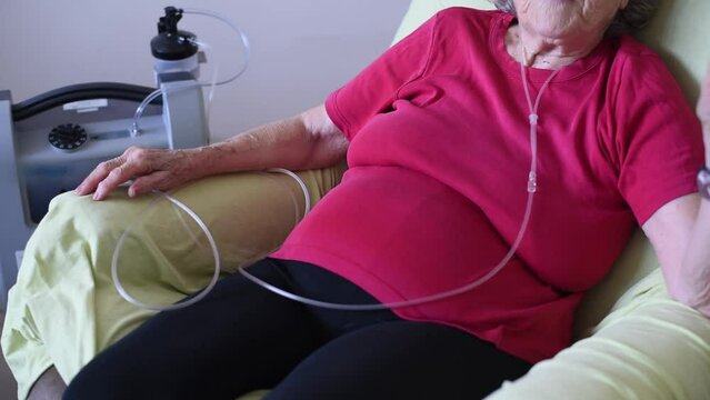 Person sit on chair using Oxygen breathing machine at home. old woman in red t-shirt. Breathing device for asthma and coronavirus. senior have problem with lungs.