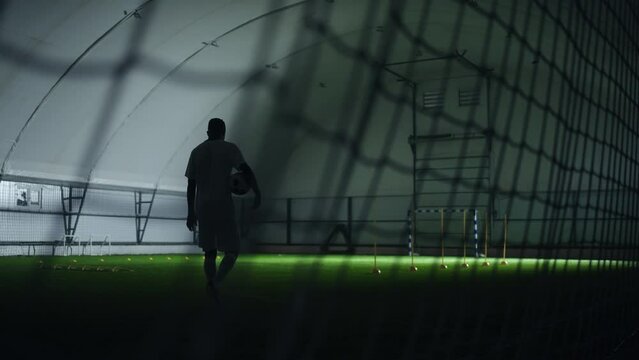 footballer in indoor training football pitch, walking with ball in hands, view through net on gates