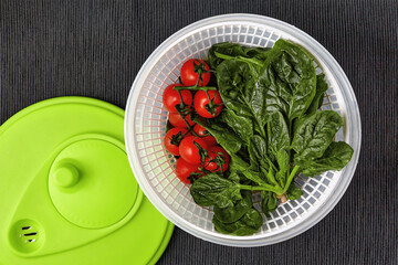 Washed red cherry tomatoes on a branch and green spinach leaves in a centrifuge strainer on a table with a black textile tablecloth