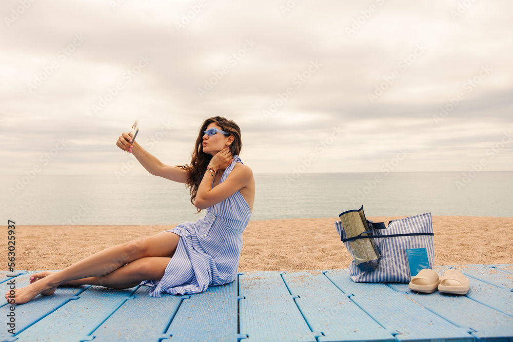 Wall mural joyful young caucasian girl sits on beach takes selfie pursing her lips in cloudy weather. brown hai