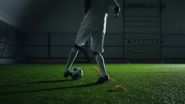 active junior sportsman training in football hall, running with ball between sticks, rear view